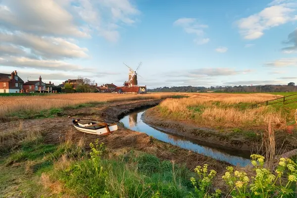 Rural Norfolk landscape with a boat in a narrow waterway, traditional windmill in the background, and houses under a partially cloudy sky.