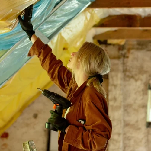 A woman installs insulation in an attic, holding a drill and adjusting a clear plastic sheet above her head.