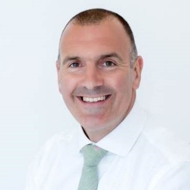 A man with short hair, wearing a white shirt and light green tie, smiles at the camera against a plain white background.