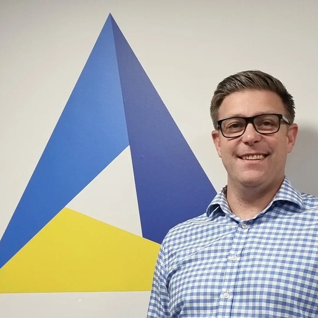 A man in glasses and a blue checkered shirt stands smiling in front of a wall with a geometric blue and yellow triangle design.