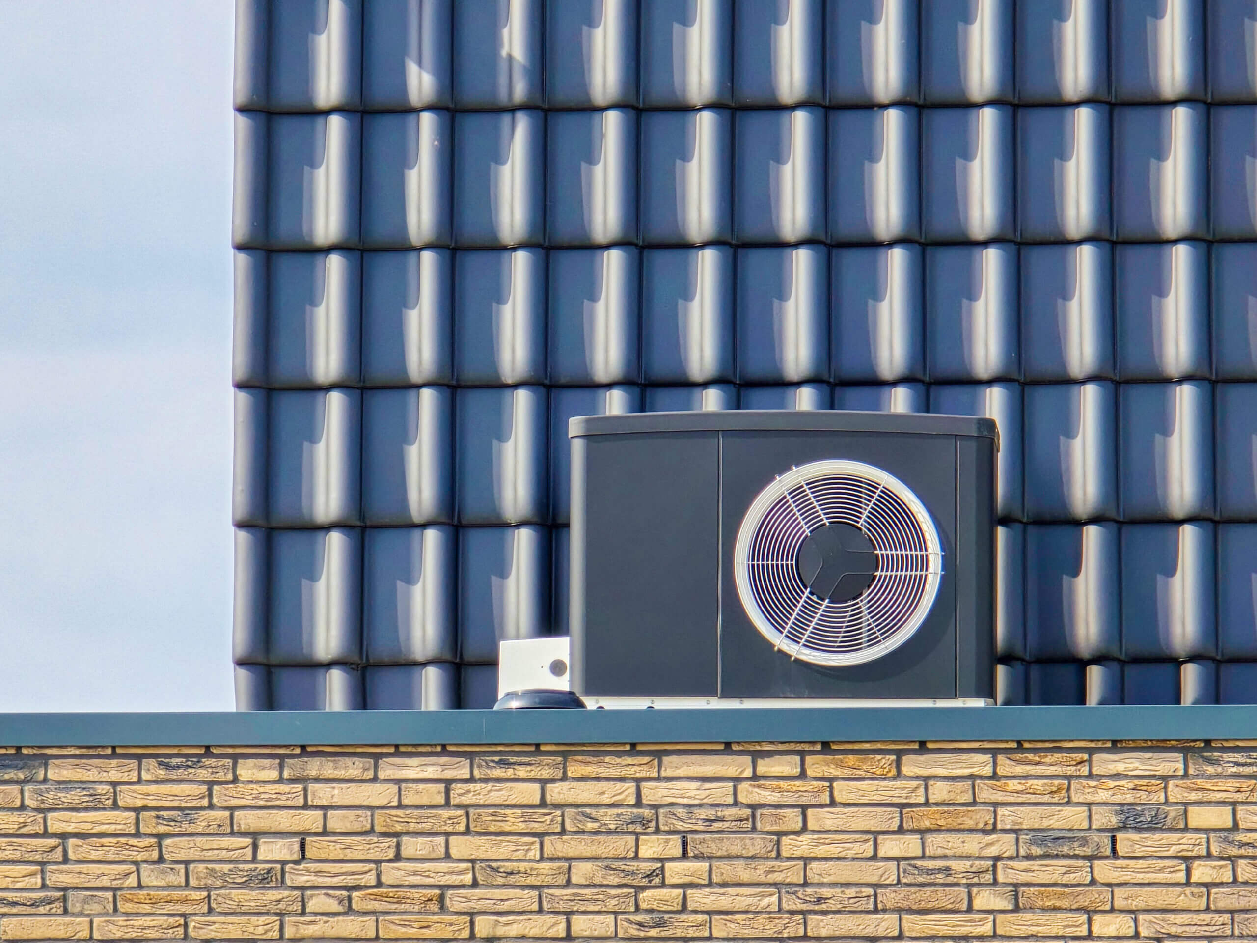 A rooftop heat pump unit sits on top of a yellow brick building, with a background of dark blue ceramic roof tiles.